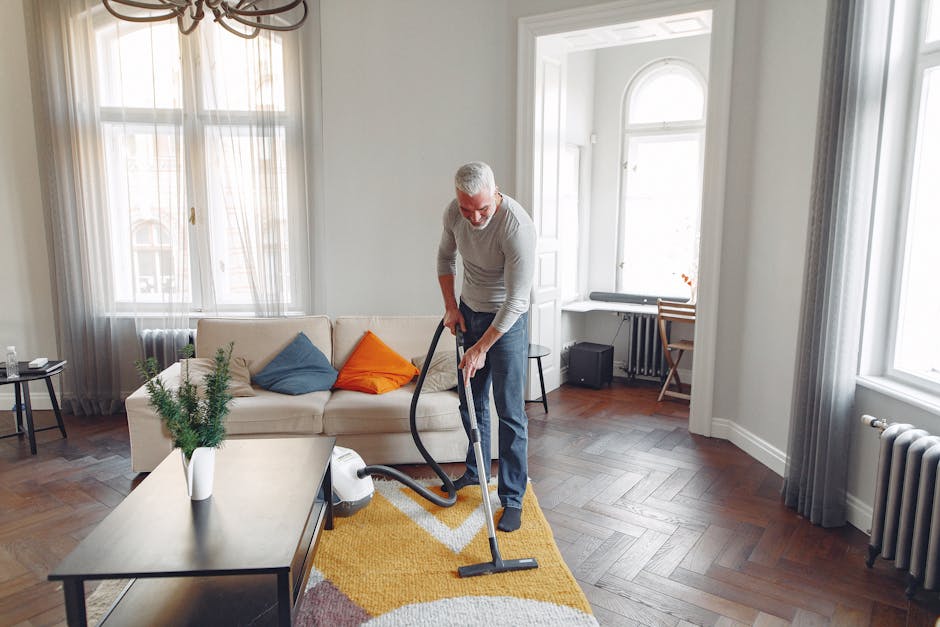 A professional cleaner in full protective gear, including a white suit, gloves, and a face mask, is using a specialized vacuum or cleaning machine to remove dust and debris from a plush, beige carpet