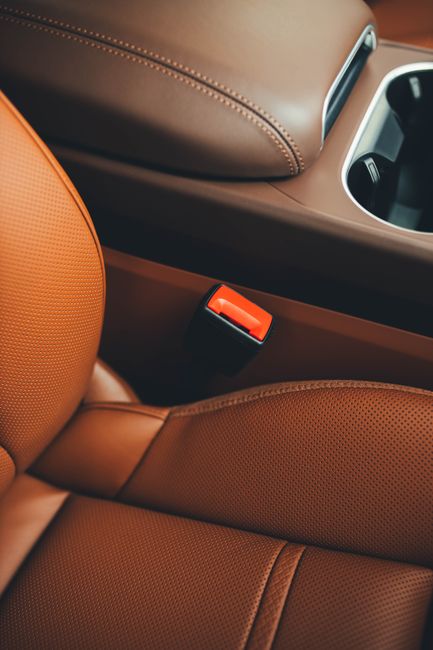 Close-up image of the interior of a vehicle, focusing on the tan leather upholstery of the passenger seat and the surrounding center console area. The seat surface shows a textured, perforated leather