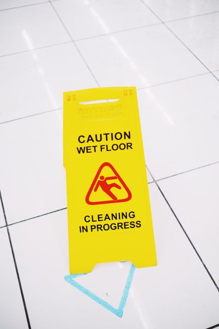 A yellow caution wet floor sign with black and red text warning of a wet surface, placed on white tiled flooring in an indoor setting. The sign indicates 'Cleaning in Progress' and features a pictogra