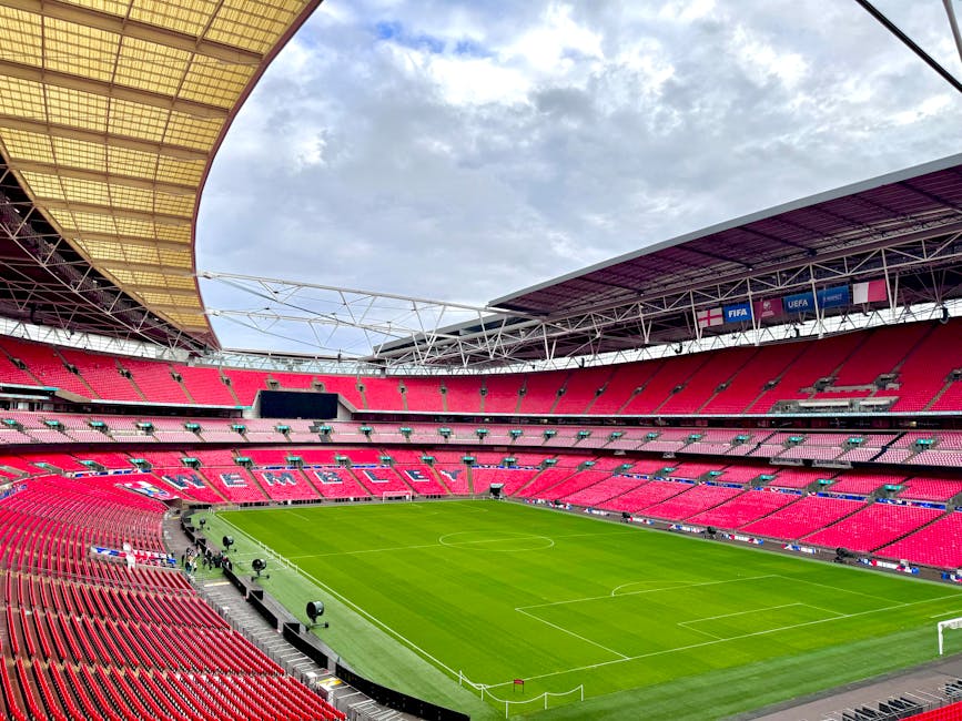 A panoramic view of Wembley Stadium's interior showcasing the soccer pitch with well-maintained green grass, marked with white lines for gameplay. Surrounding the field, the vibrant red spectator seat